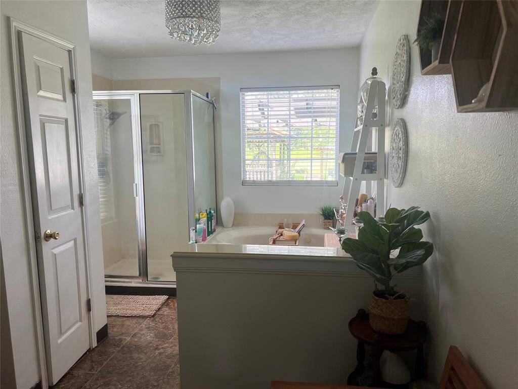 This is a bright, well-lit master bathroom featuring a combination of a glass-enclosed shower and a separate bathtub. The room includes a window with blinds, elegant light fixture, and decorative shelving, creating a cozy and inviting space.