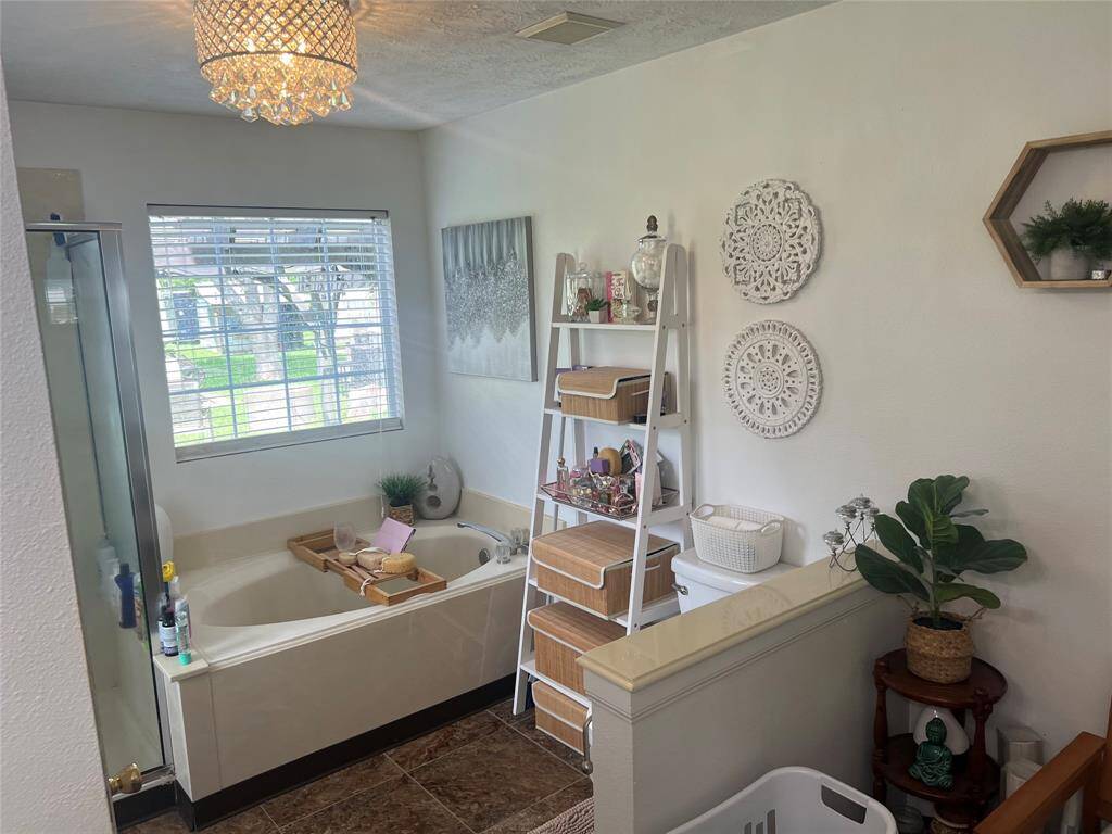 This is a cozy, well-lit master bathroom featuring a large tub with a shower option, decorative tile flooring, and a window that provides natural light. The room is accessorized with an elegant chandelier, decorative wall art, and a shelving unit for storage, creating a comfortable and stylish space.