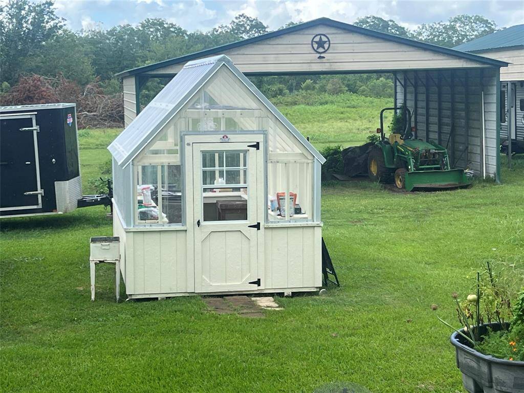 You're looking at a charming garden shed with clear windows and a peaked roof, set in a lush, green yard. Adjacent to the shed is an open-sided metal carport with a tractor parked underneath, indicating ample storage and workspaces on the property.