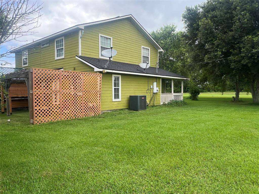 This is a two-story, yellow house with white trim, featuring a covered porch, satellite dish, and an outdoor A/C unit. It has a spacious, well-maintained lawn with mature trees in the background. A wooden lattice structure is present on the left, possibly for privacy or as a trellis.