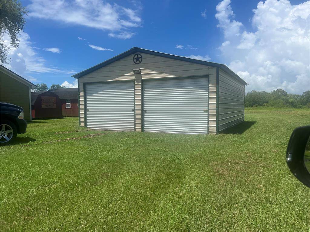 This is a photo of a standalone metal garage or storage shed with a gable roof and dual sliding doors, situated next to three car garage with ample green lawn space.