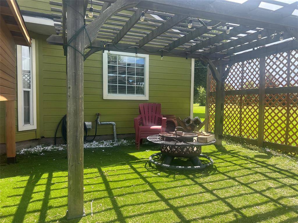 A cozy backyard space with a green lawn, featuring a pergola providing partial shade, comfortable outdoor seating, and a fire pit table, ideal for relaxation and entertainment. The wooden lattice adds privacy and the exterior of the house is finished in an inviting green siding with white trim around the window.