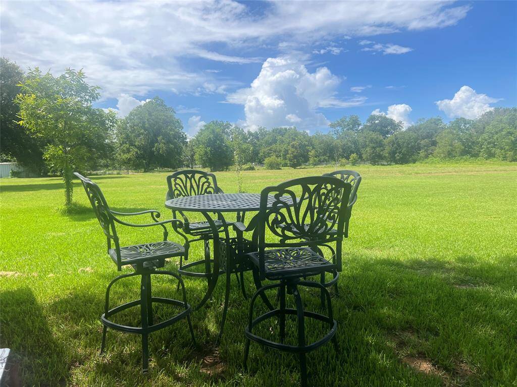 A tranquil outdoor setting with a metal patio table and chairs on a lush green lawn, surrounded by trees and an expansive grassy area under a sunny sky with some clouds. Ideal for those seeking a peaceful backyard retreat.