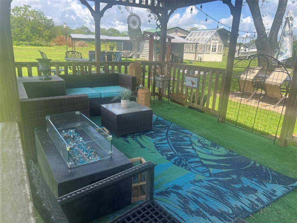 A cozy outdoor patio space with comfortable lounge furniture, a decorative rug, and a fire pit table, all enclosed by a wooden fence with a view of a green lawn and surrounding structures, offering a private and inviting area for relaxation and entertainment.