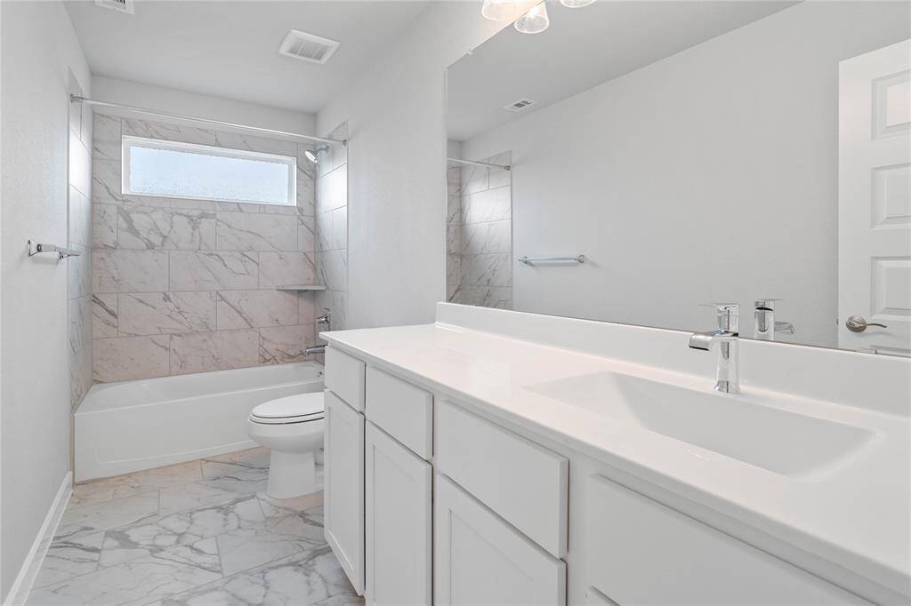 Secondary bath features tile flooring, bath/shower combo with tile surround, white stained wood cabinets, beautiful light countertops, mirror, dark, sleek fixtures and modern finishes.