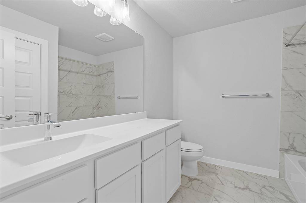 Secondary bath features tile flooring, bath/shower combo with tile surround, white stained wood cabinets, beautiful light countertops, mirror, dark, sleek fixtures and modern finishes.