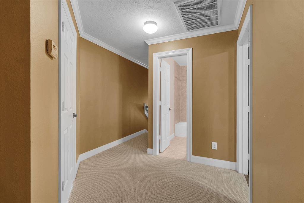 This is hallway with neutral-toned walls, textured carpet, and white trim around the doors. The hallway leads to a bathroom, and there are multiple doors for room access. A central light fixture provides illumination.