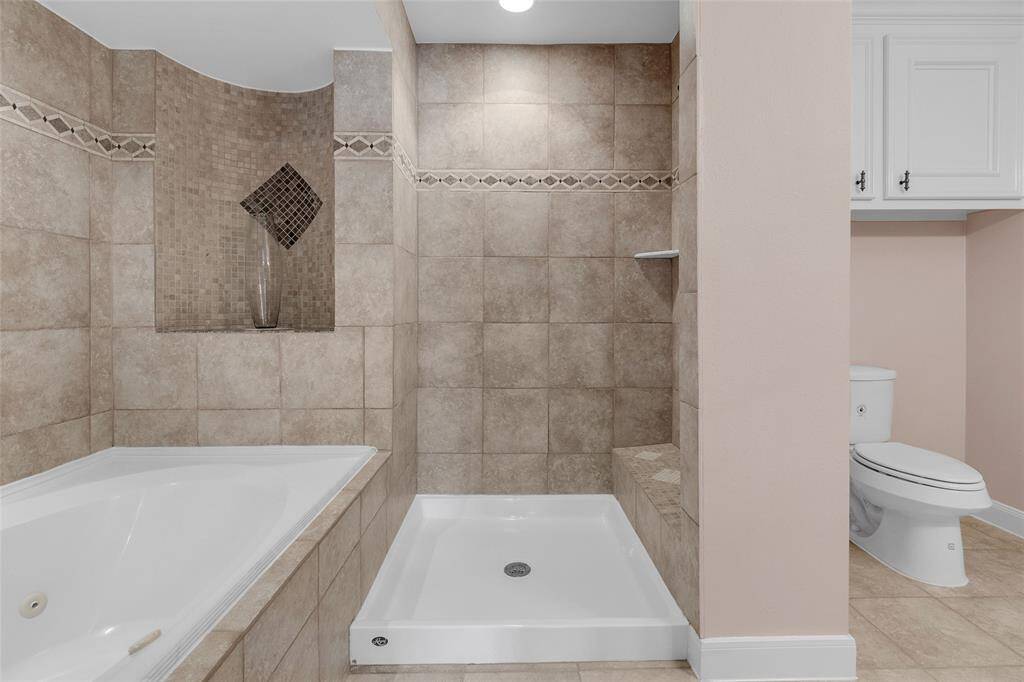 Modern bathroom with a corner soaking tub, a separate shower stall, and a toilet. Neutral tiles with decorative accents line the walls, and there's built-in cabinetry for storage.