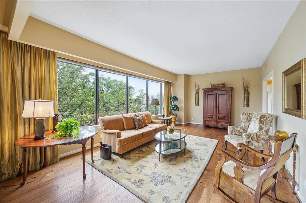 Bright living room with large windows offering a view of trees, featuring hardwood floors, a cozy sofa, elegant armchairs, and a classic armoire.