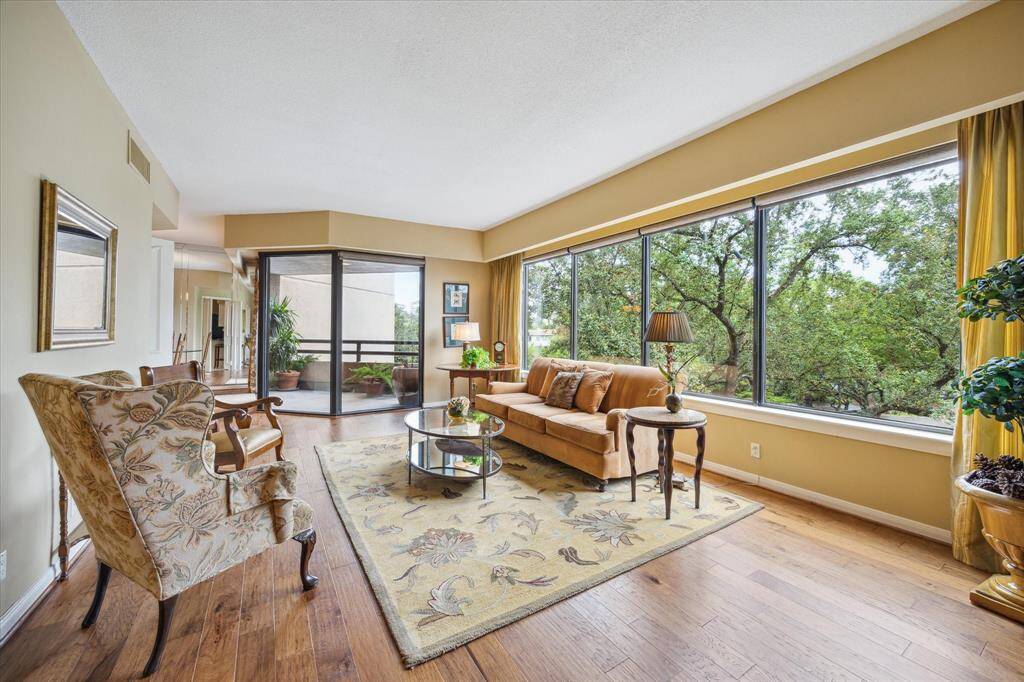 This bright living room features large windows with lush green views, hardwood floors, and cozy seating. Adjacent is a balcony with potted plants, enhancing the indoor-outdoor connection. Perfect for a relaxing, nature-inspired atmosphere.