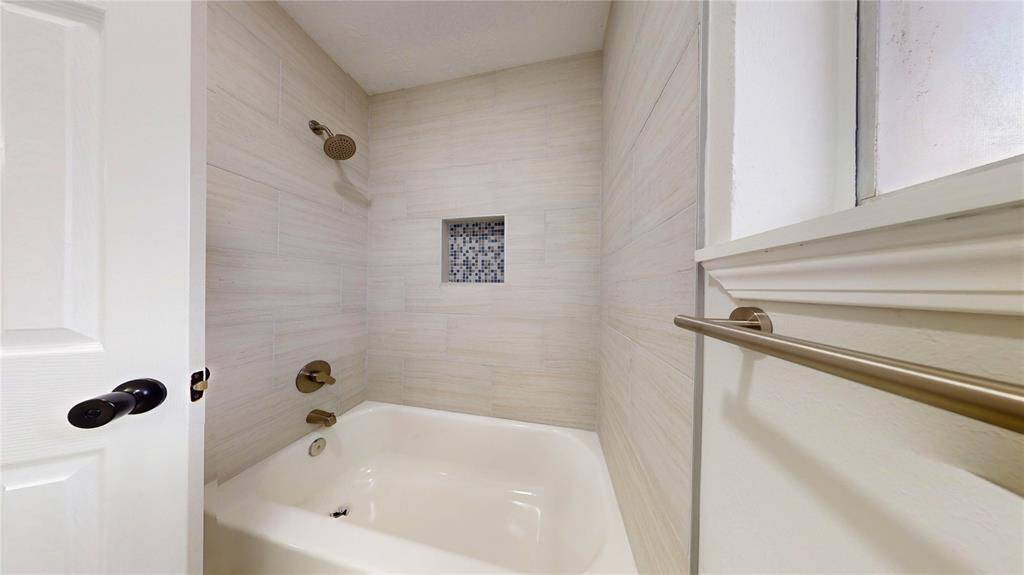 White bathtub with an overhead shower, textured wall tiles, and a small decorative tile niche. There's a window providing natural light.