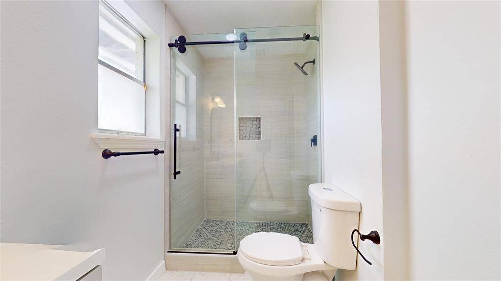 Bathroom featuring a glass-door walk-in shower with tile surround, a standard toilet, and a window for natural light.