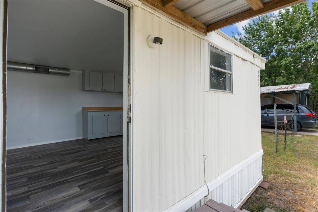 This photo showcases the exterior of a mobile home with a partially open door revealing a glimpse inside. The interior features wood-style flooring and cabinetry. Outside, there's a small covered porch, some greenery, and a carport with a vehicle parked.