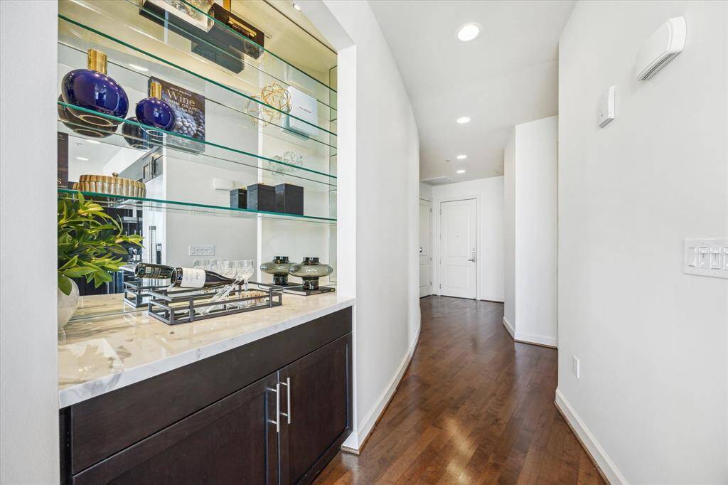 This photo showcases a sleek, modern hallway with polished wood flooring and recessed lighting. On the left, there's a built-in display with glass shelves and a marble countertop, perfect for showcasing decorative items or a mini bar. The space feels open and inviting, leading towards a door at the end.