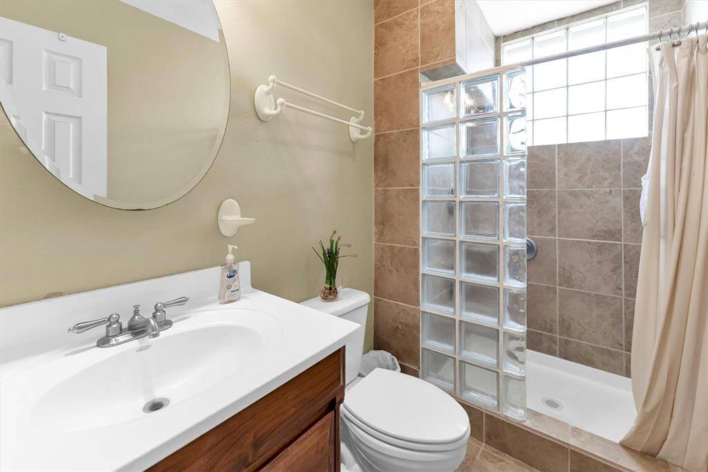 This bathroom features a single sink vanity with a wood finish, a large circular mirror, and a tiled shower-tub combo with glass block windows for privacy and natural light, and is equipped with grab bars.