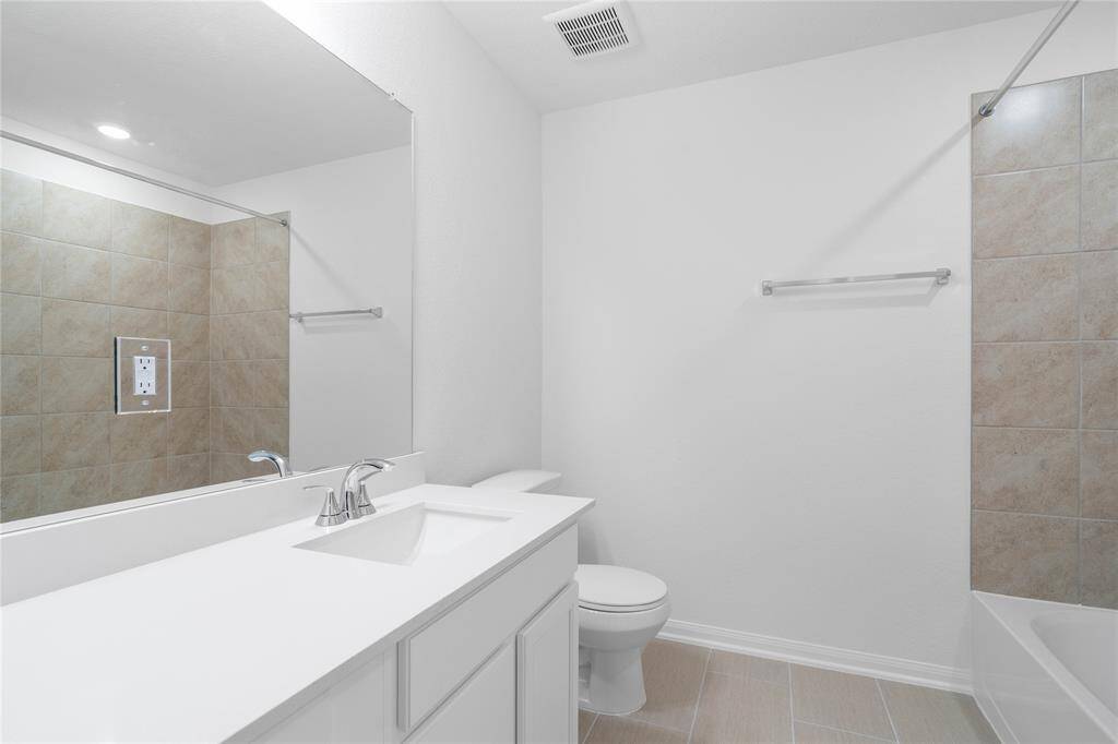 Secondary bath features tile flooring, bath/shower combo with tile surround, stained wood cabinets, beautiful light countertops, mirror, dark, sleek fixtures and modern finishes.