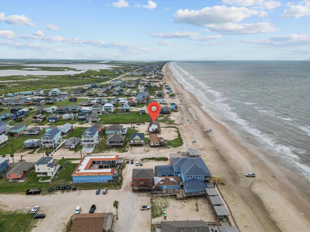 This aerial photo showcases a beachside neighborhood with a house marked by a red pin. The property is directly facing the ocean, offering immediate beach access and scenic views. Nearby houses are spaced with green lawns, providing a relaxed coastal community feel.