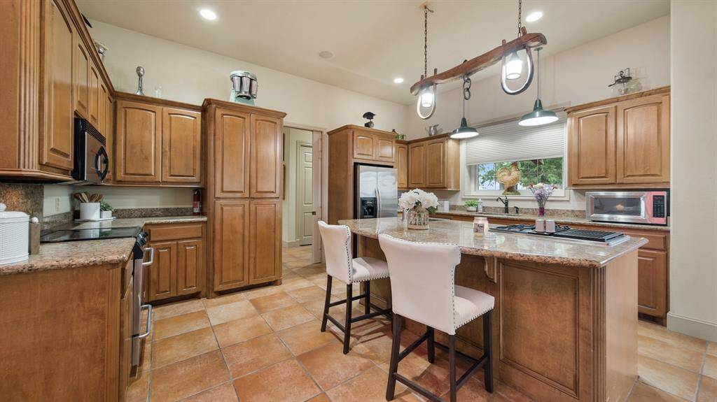 This is a spacious kitchen with ample wood cabinetry, granite countertops, modern appliances, a central island with bar seating, and attractive pendant lighting, set on a tiled floor.