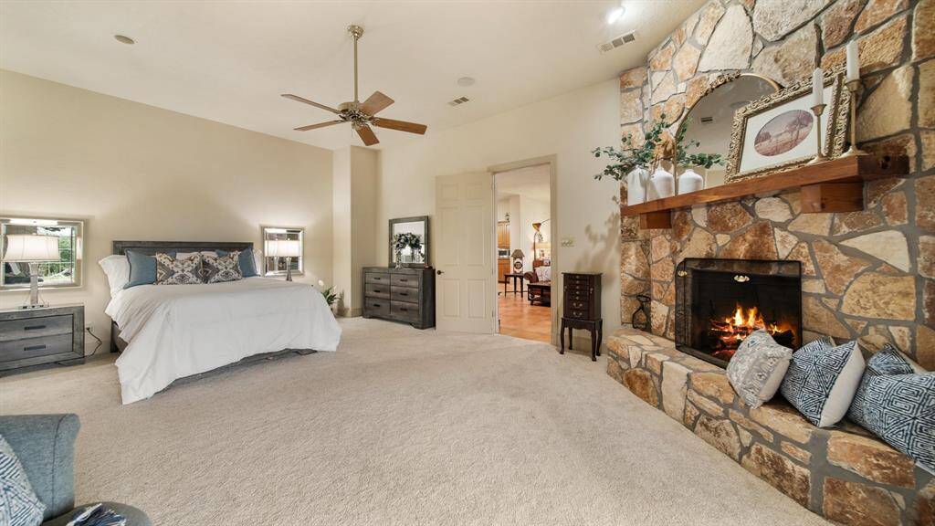 This roomie primary bedroom features a fireplace with stone mantle, plush carpeting, a ceiling fan, and a soothing neutral color palette. It offers