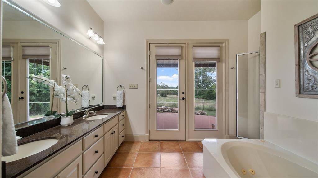 This is a spacious Primary bathroom with a dual-vanity sink, large mirror, tiled floor, and a separate jetted bathtub and shower. It features ample natural light and has a double French door.