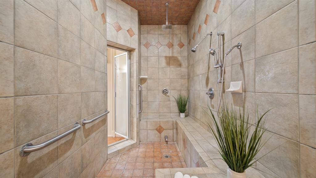 This is a spacious walk-in shower with beige tile walls, accented with decorative diamond-shaped tiles, featuring multiple shower heads and built-in shelving, complete with accessibility handles.