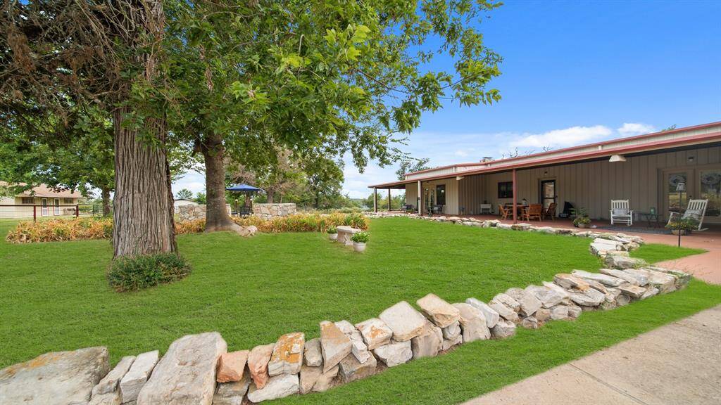This is a single-story Barndo style home with a spacious, mature trees and a stone sitting area. The exterior includes a covered patio area perfect for outdoor relaxation.