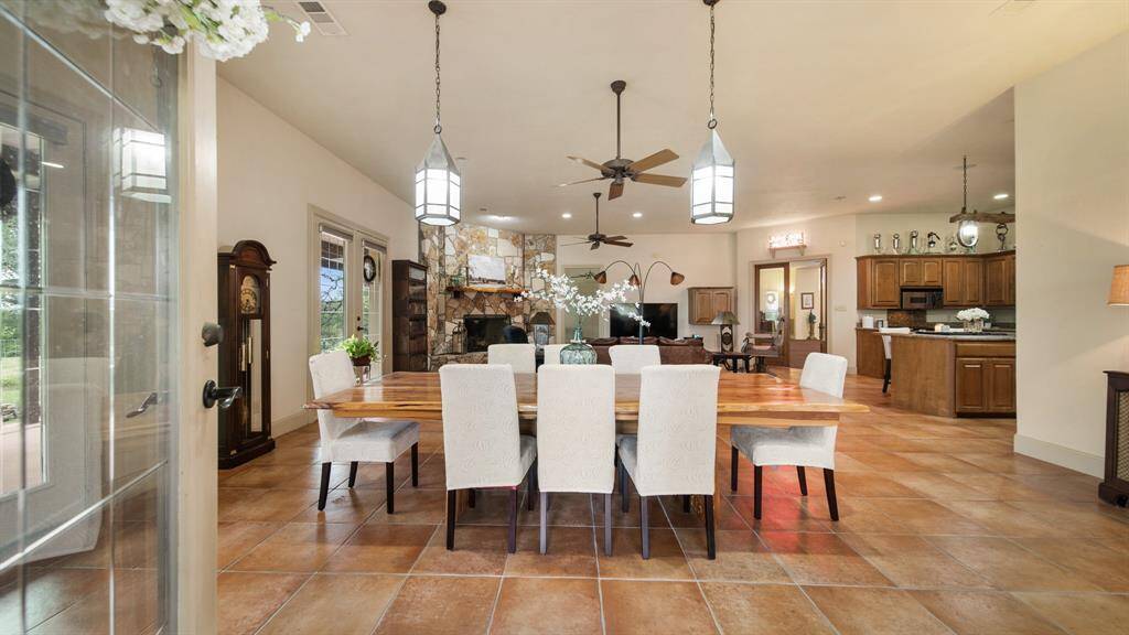 This is a spacious, open-concept dining area with an adjoining kitchen, featuring terracotta tiled floors, elegant lighting fixtures, and a warm, inviting color scheme. The room is well-lit with natural light from your walls of French doors
