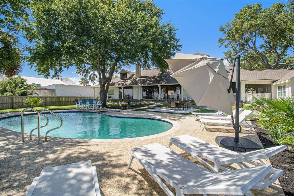This photo showcases a spacious backyard with a well-maintained in-ground pool, surrounded by mature trees for privacy. Ample lounging space, a patio area with outdoor dining, and a sunny ambiance make it ideal for relaxation and entertainment.