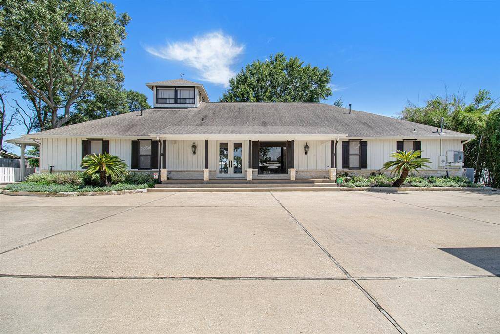 This is a spacious single-family home with a large driveway, featuring a symmetrical façade with a central entrance, flanked by windows and greenery. The house has a two-story section with a prominent window, and a single-story wing with a covered porch area.