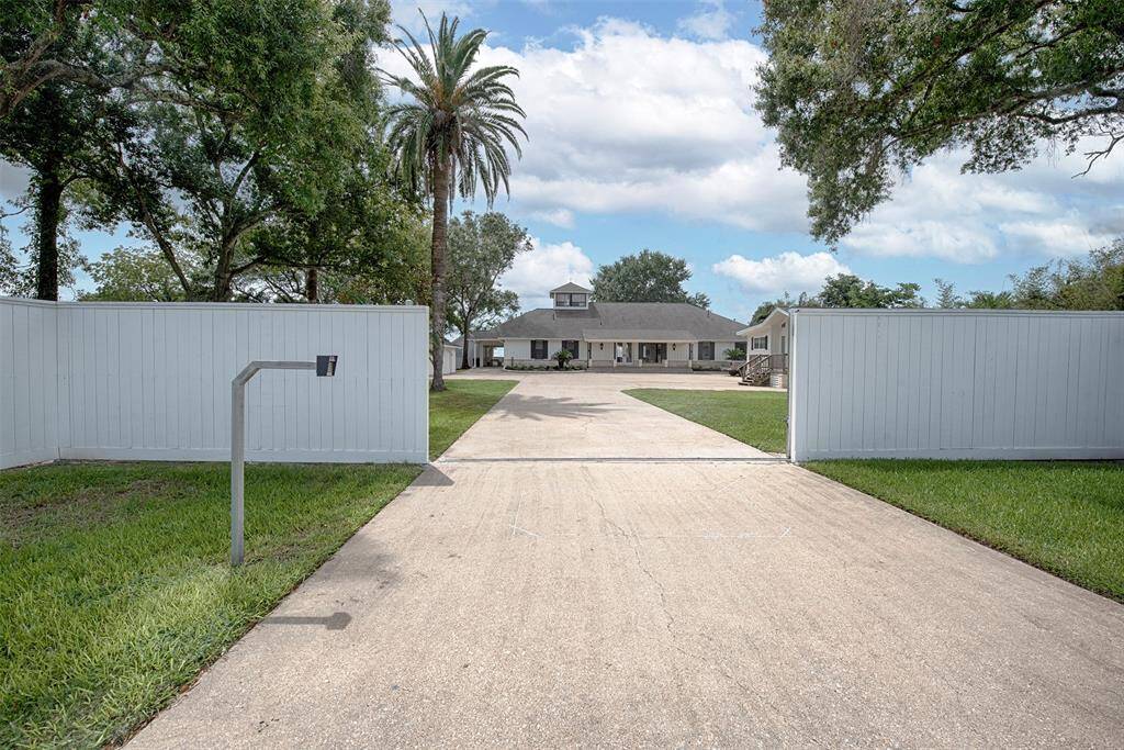 Gated property featuring a long driveway that leads to a two-story house with a prominent front staircase. The home is flanked by mature trees and a tall privacy fence. The setting suggests a private, suburban feel.