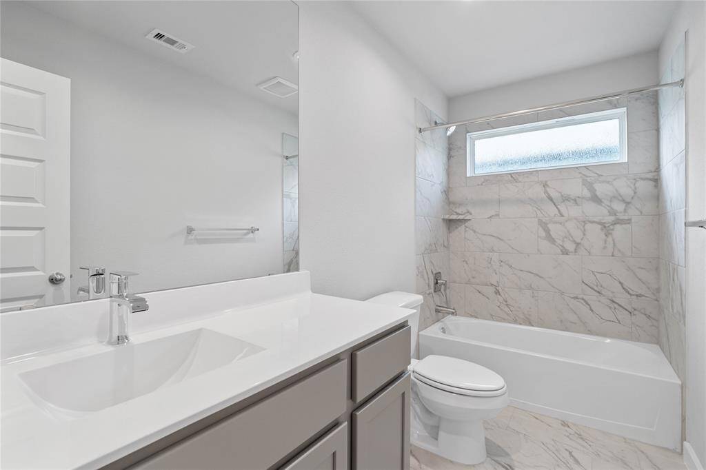 Secondary bath features tile flooring, bath/shower combo with tile surround, stained wood cabinets, beautiful light countertops, mirror, dark, sleek fixtures and modern finishes.