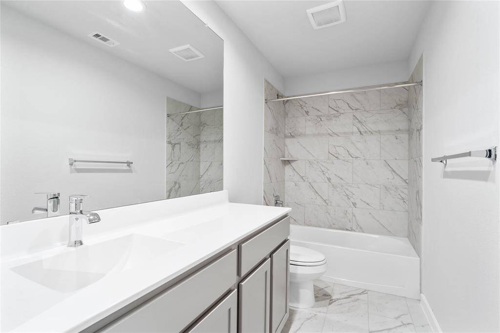 Secondary bath features tile flooring, bath/shower combo with tile surround, stained wood cabinets, beautiful light countertops, mirror, dark, sleek fixtures and modern finishes.