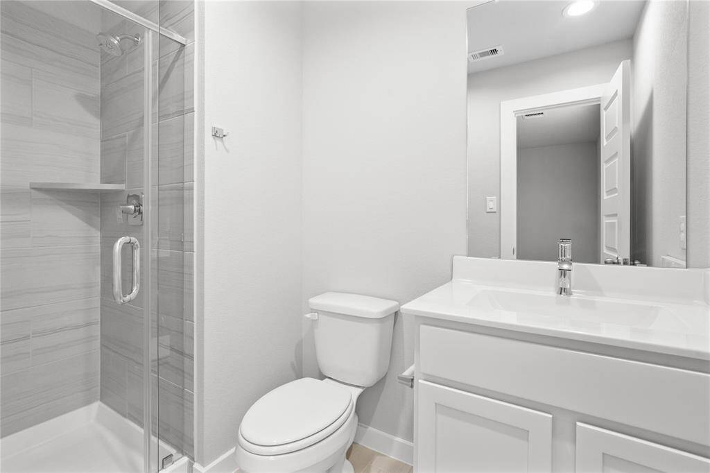This private bath features tile flooring, shower with tile surround, light stained wood cabinets, beautiful light countertops, mirror, dark, sleek fixtures and modern finishes.