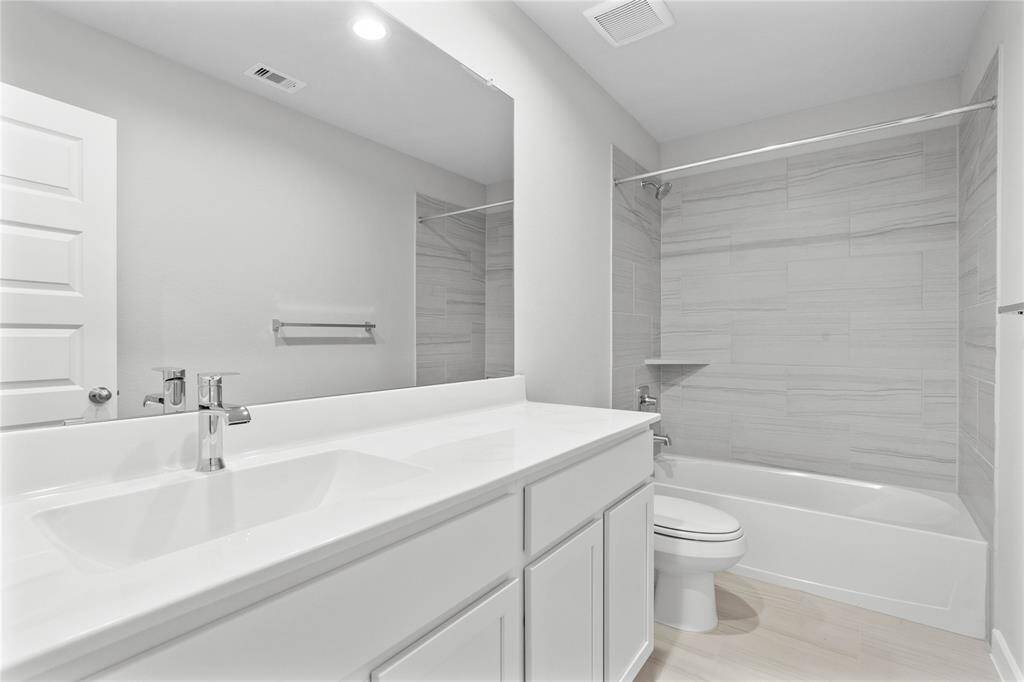 Secondary bath features tile flooring, bath/shower combo with tile surround, stained wood cabinets, beautiful light countertops, mirror, dark, sleek fixtures and modern finishes.