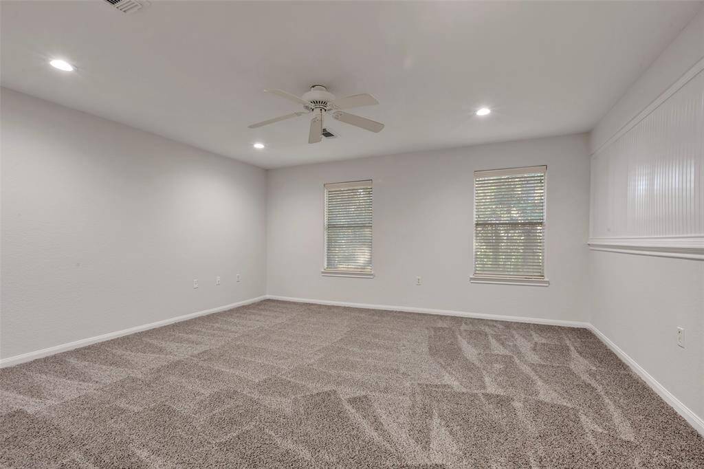 This is a spacious, well-lit room featuring plush carpeting, two windows with horizontal blinds, a ceiling fan, and recessed lighting. It has neutral-colored walls and ample wall space for customization.