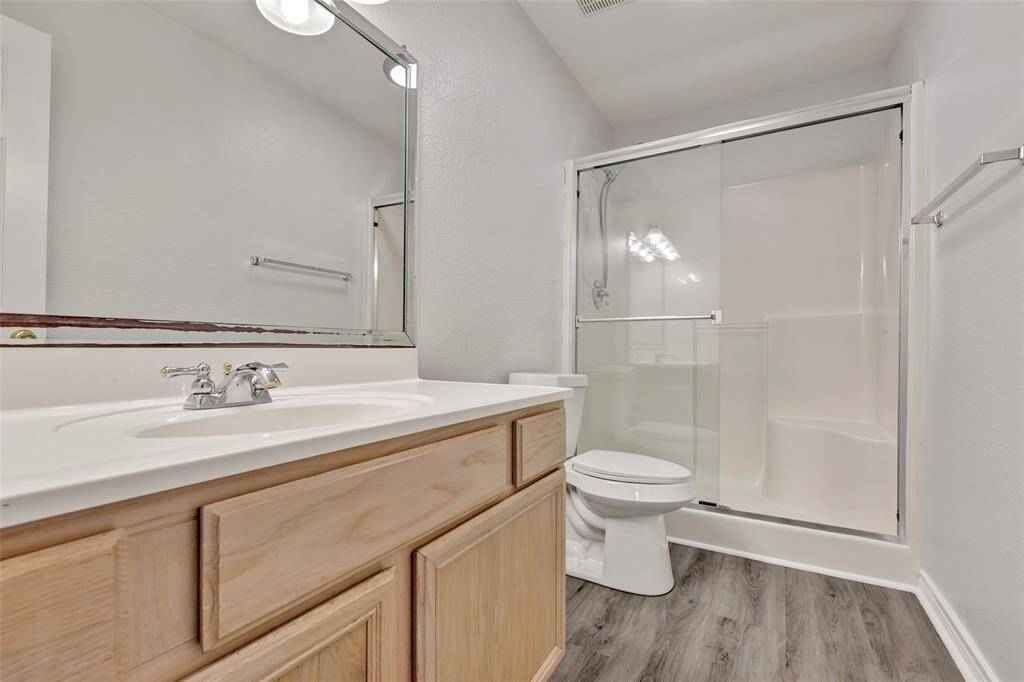 This is a clean and modern bathroom with vinyl plank flooring, a large mirror over a single sink vanity, a toilet, and a full-size shower with sliding glass doors.