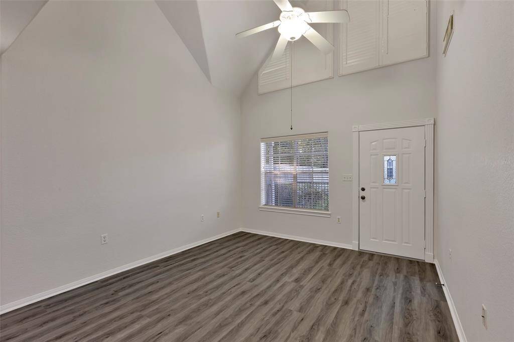 This is a bright, spacious living room with high vaulted ceilings, new vinyl plank flooring, a large window for natural light, and a ceiling fan. The entry door features decorative glass detailing.
