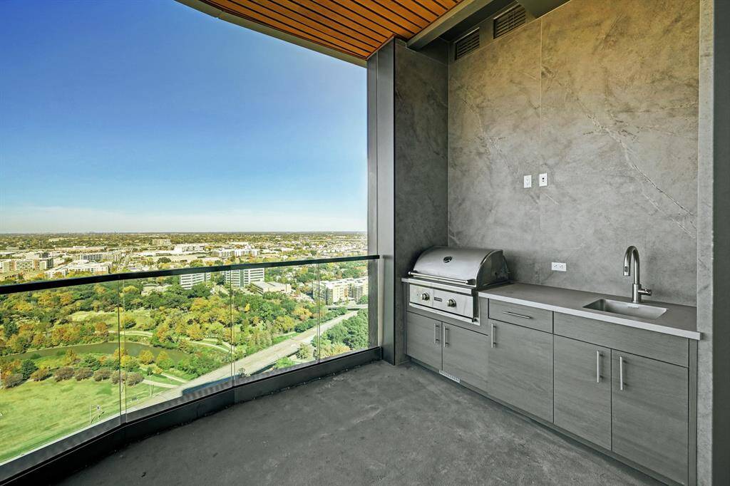 This photo showcases an outdoor kitchenette on a high-rise balcony with a built-in grill and sink. It is set against a breathtaking, expansive view of the cityscape and parkland.