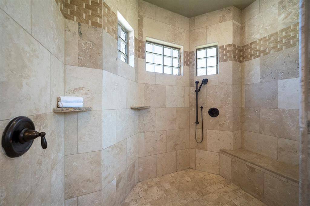 This is a spacious walk-in shower with neutral-toned tile work, featuring a built-in bench, a handheld showerhead, and natural light from two frosted glass windows.