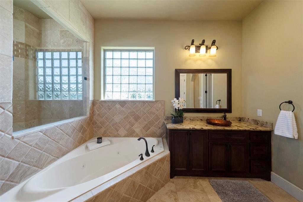This is a spacious and well-lit bathroom featuring a large corner bathtub with a tiled surround, a glass block window for privacy and natural light, a dark wood vanity with a granite countertop and vessel sink, and a modern light fixture above a framed mirror. The room is finished with neutral tones and a tiled floor.