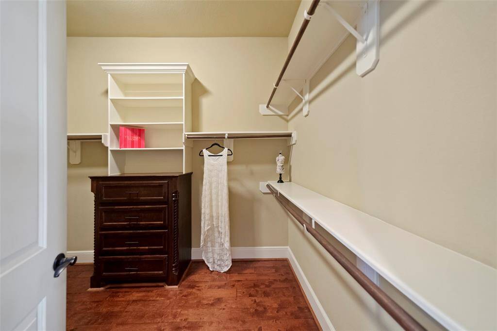 This is a spacious walk-in closet with built-in shelves, ample hanging rods, and a dresser for additional storage. The room features warm-toned flooring and neutral wall colors, offering a clean and organized space for clothing and accessories.