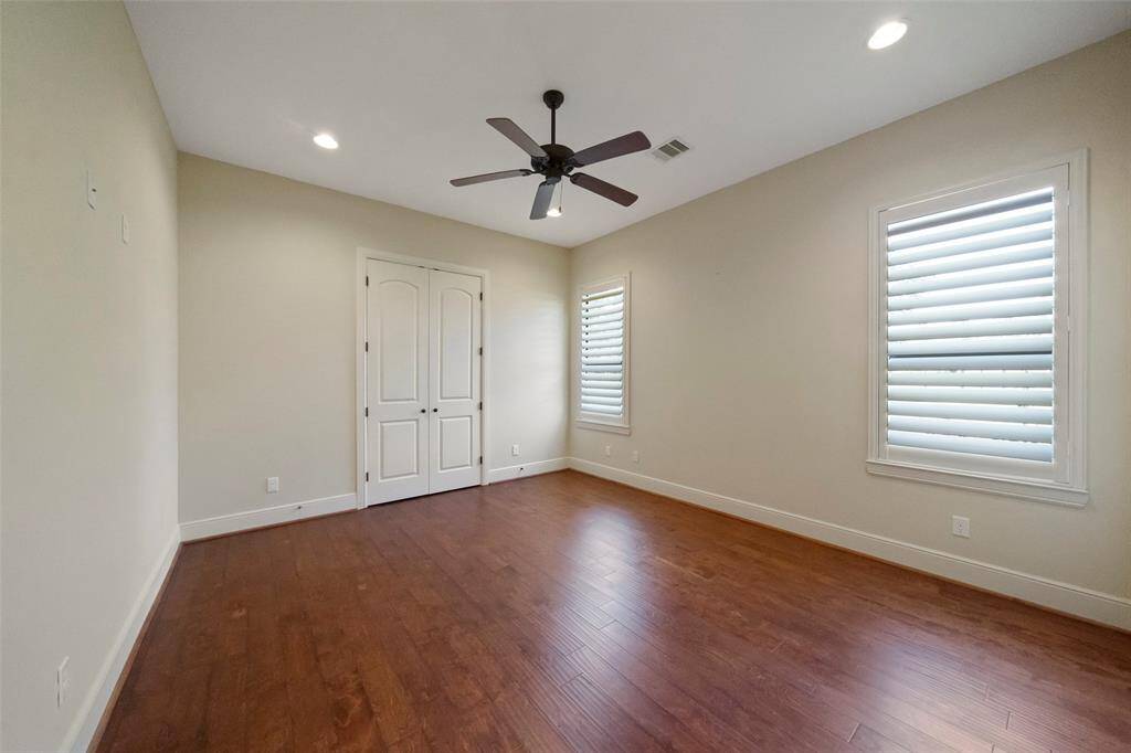 This is a spacious, well-lit bedroom featuring hardwood floors, a modern ceiling fan, and large windows with plantation shutters. It has a clean, neutral color scheme and includes a double-door closet for storage.