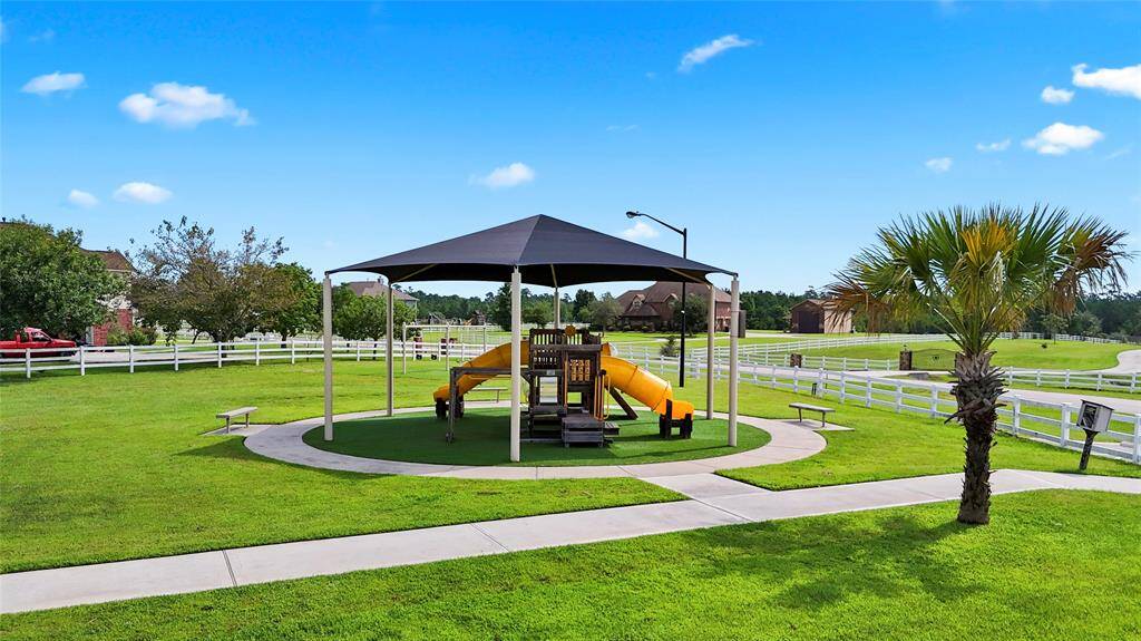 This photo features a well-maintained community playground with a shaded play structure, benches, and lush green lawn, surrounded by a white fence with residential homes in the background. It's an ideal outdoor space for families.