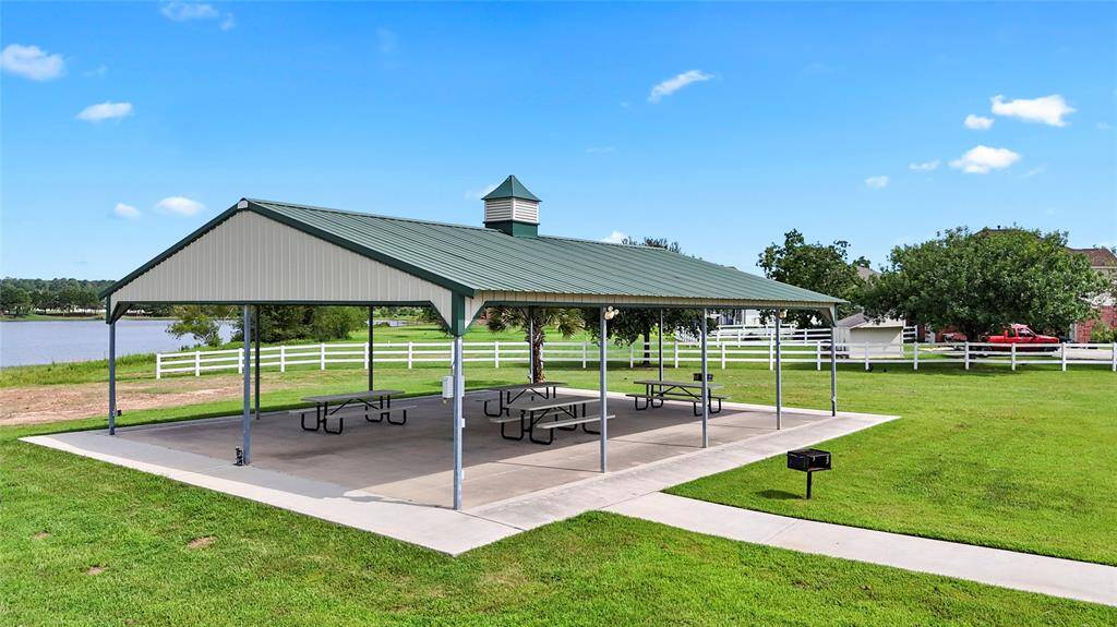 This is a community park area with a large covered pavilion featuring picnic tables and a barbecue grill, set against a scenic backdrop with a lake and neatly maintained lawns. A perfect spot for outdoor gatherings and family picnics.