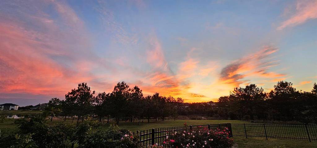 This property offers a serene backyard view with a picturesque sunset sky, complementing the peaceful landscape with lush greenery and flowering shrubs, all enclosed by an elegant black fence.