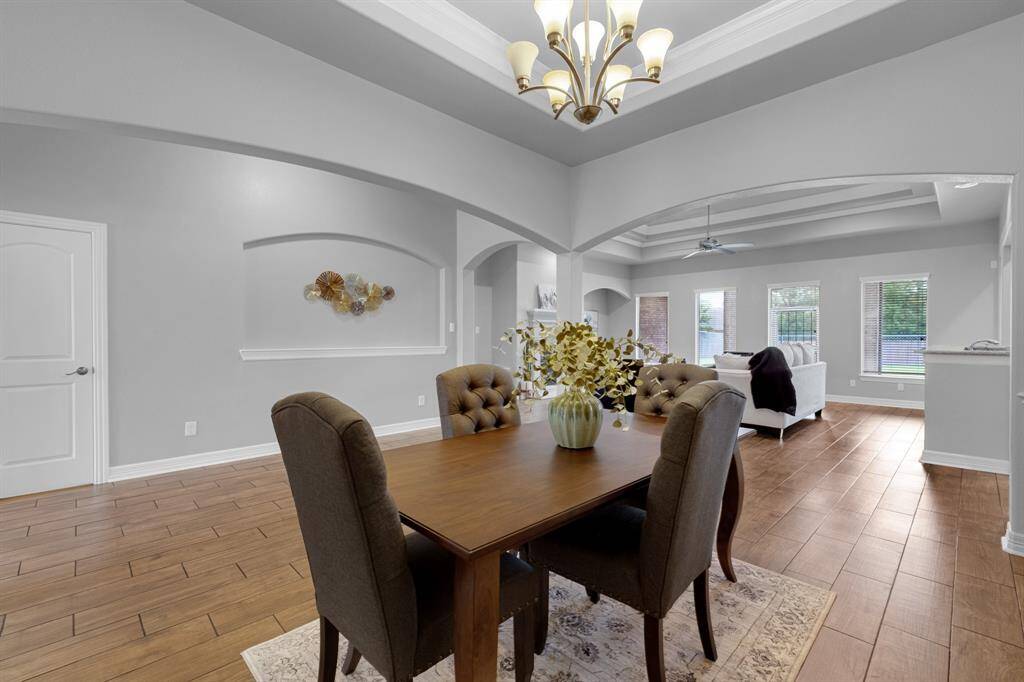 This is a spacious, open-concept dining area with elegant tray ceilings and modern lighting, leading into a cozy living space with abundant natural light from large windows. The room features hardwood flooring and neutral color tones, offering a blend of sophistication and comfort.