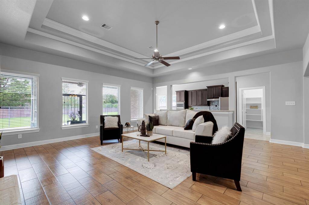 This is a spacious, modern living room with high coffered ceilings, large windows providing ample natural light, elegant hardwood flooring, and a neutral color palette. The open layout offers a seamless flow into the adjoining kitchen area.