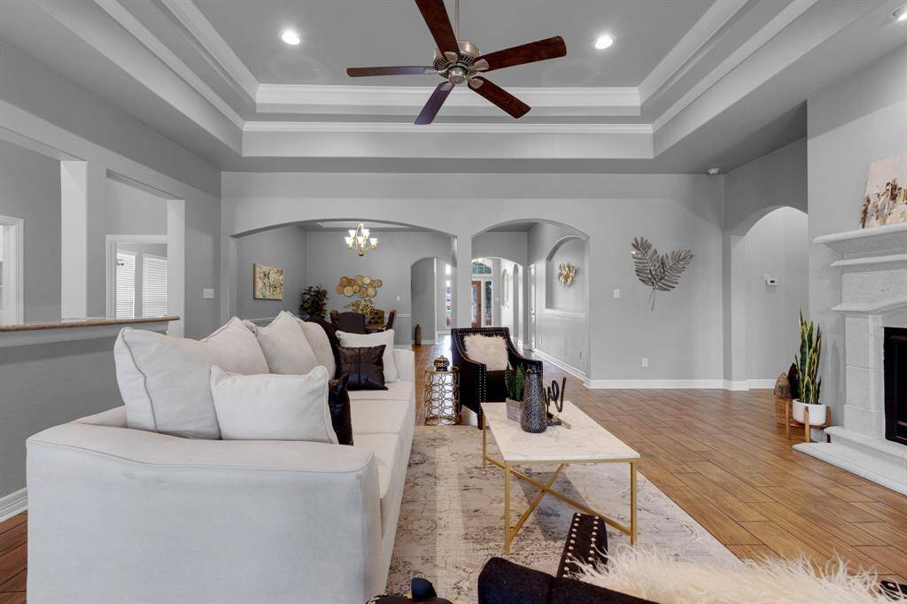 This is a spacious and elegantly designed living room featuring high ceilings with crown molding, a cozy fireplace, hardwood floors, and arched walkways that lead to other parts of the home. The room is well-lit and includes a ceiling fan, with a neutral color palette that offers a modern and sophisticated feel.