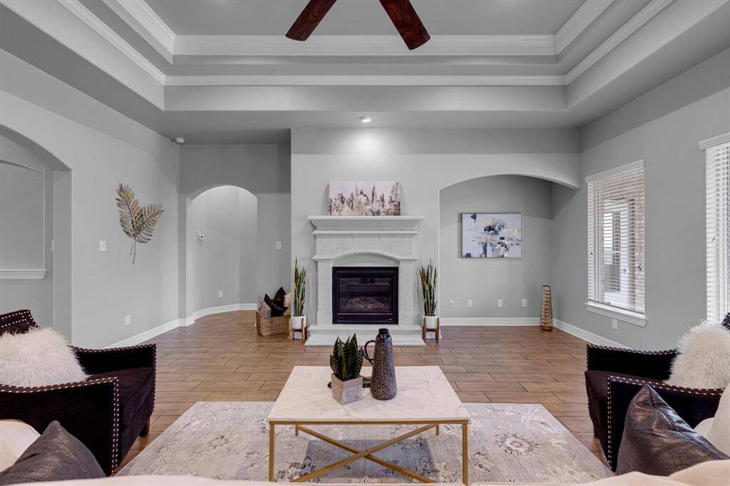 This is a spacious living room with high ceilings, featuring crown molding and exposed wooden beams. It includes a fireplace as a centerpiece, arched walkways, and ample natural light from large windows. The room is finished with a neutral color palette and hardwood flooring.