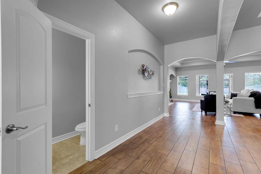 This is a spacious and well-lit interior with an open floor plan featuring archways, recessed lighting, and hardwood floors. There is a view into a cozy living room with large windows and a glimpse of a bathroom to the side.